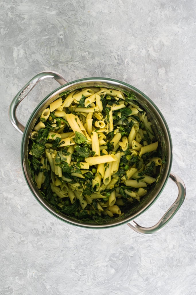 A large saucepan filled with cooked penne pasta tossed with spinach, peas, nutritional yeast, turmeric apple cider vinegar and healthy avocado oil.