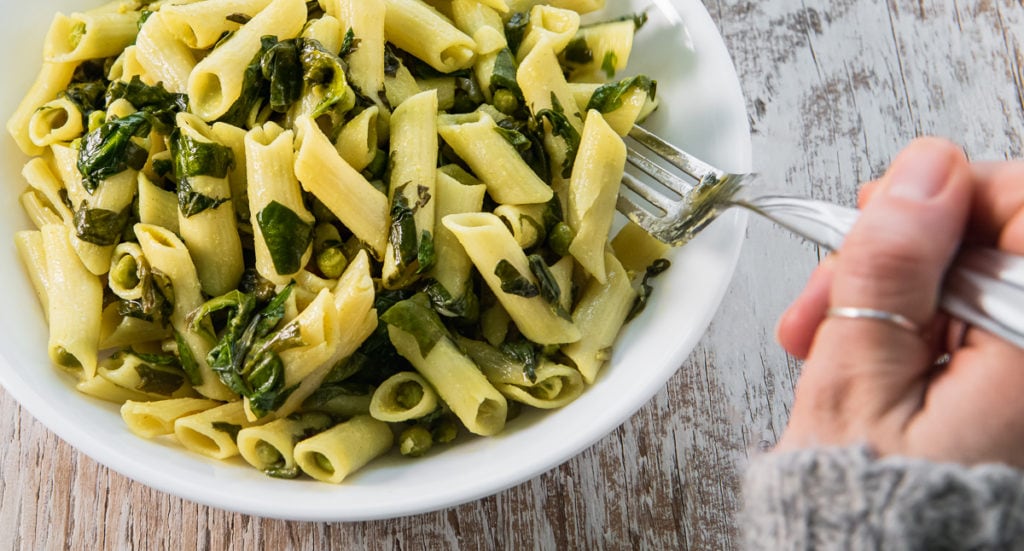 A hand holding a fork, digging into a big bowl of spinach and peas penne pasta.