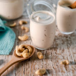 A little cup of creamy cashew yogurt, swirled to the top with another cup full and a larger jar behind, as well as a spoon of cashews spilling about.
