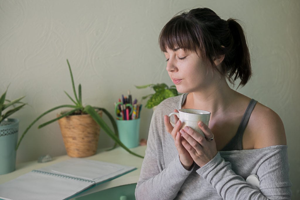 A young woman sitting at a desk with plants, a pen holder and an open notebook, holding a mug close with both hands and her eyes closes.