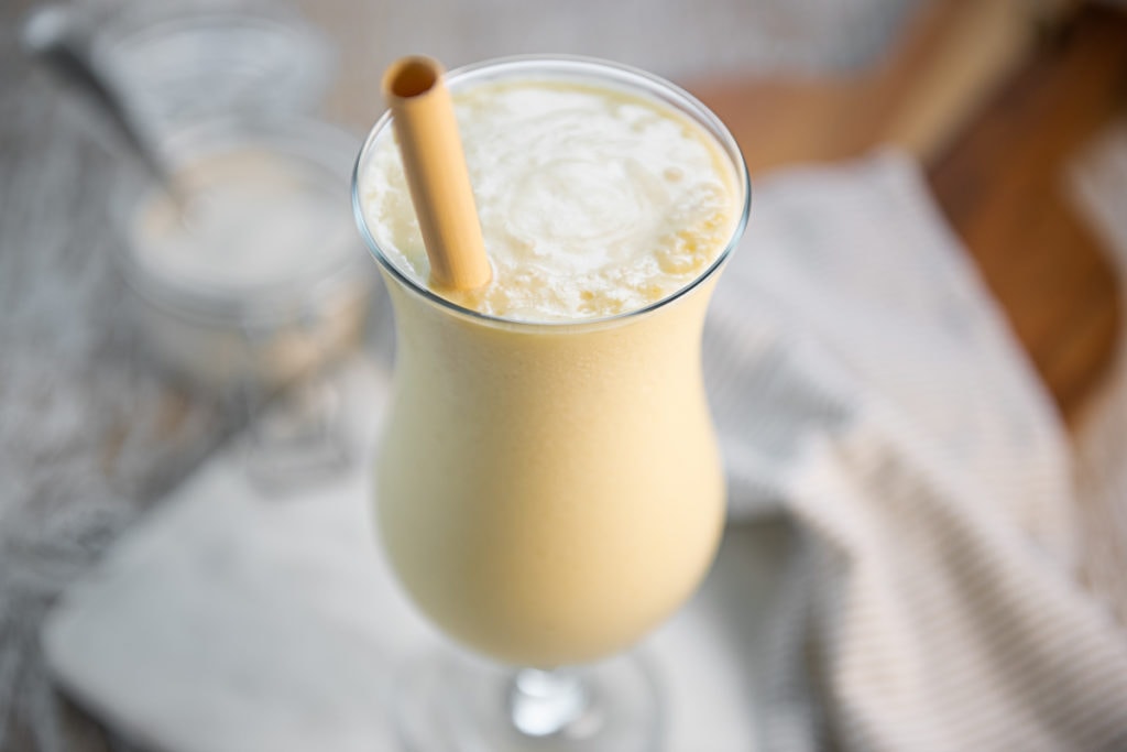 A refreshing, yellow Pineapple Smoothie in a Poco Grande Glass on a white marble cutting board with a linen cloth and a jar of yogurt in the background.