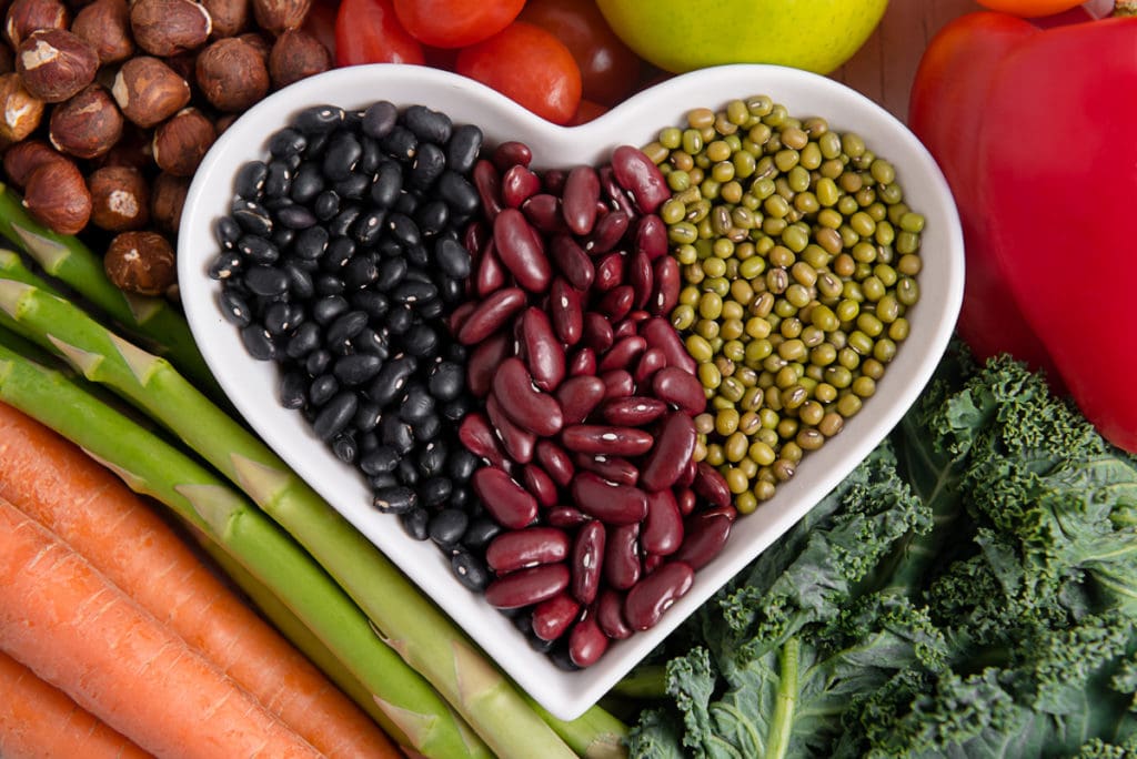 A heart shaped bowl holding three different kinds of beans, surrounded but fresh vegetables.