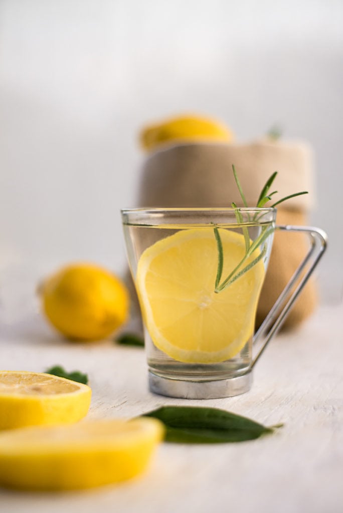 A clear mug with water, lemon and a sprig of rosemary,