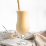 A chilled, soft yellow Pineapple Smoothie in a Poco Grande (AKA Pina Colada) Glass, sitting sitting onto of a marble cutting board with a napkin and a jar of yogurt in the background.