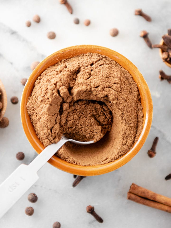 A measuring spoon dipped in a bowl of pumpkin spice with cloves, allspice and cinnamon sticks dropped all around.