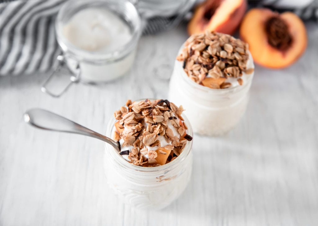 Two heaping jars of Vegan Peaches and Cream topped with skillet granola, sitting in front of an open jar and a freshly sliced peach.
