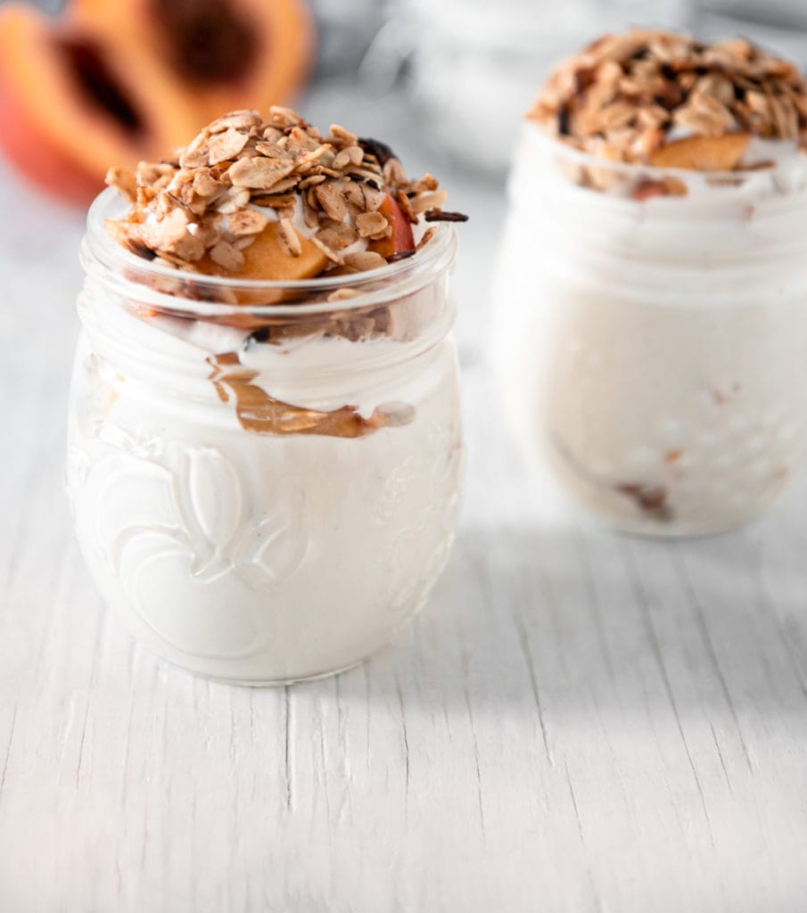 Two round jam jars filled with cashew cream fresh peaches, topped with crunchy golden granola. Sitting on a white table top with fresh peaches, a jar and a linen cloth in the background