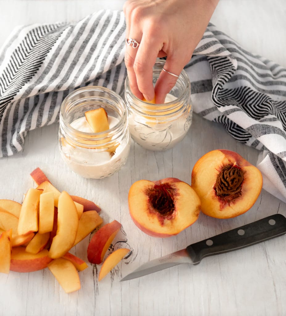 A peach sliced in half beside a pile of peach slices, with a hand placing the peaches slices into a jar of cream.