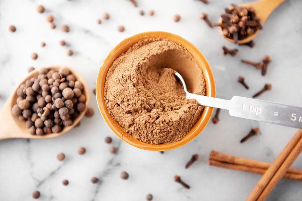 A measuring spoon dipped in a bowl of pumpkin spice with cloves, allspice and cinnamon sticks dropped all around.