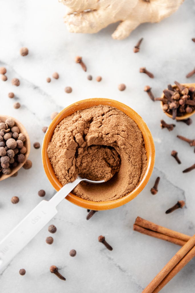 A measuring spoon dipped in a bowl of pumpkin spice with cloves, allspice and cinnamon sticks dropped all around.