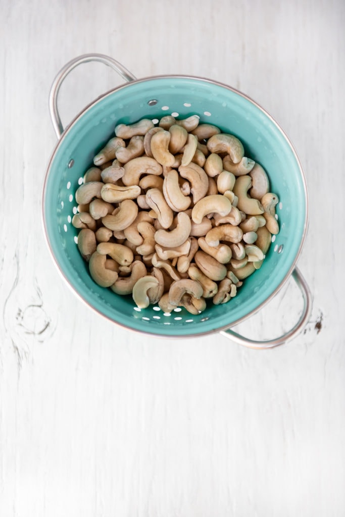 Soaked and rinsed cashews in a aqua-coloured colandar on a white wooden table top.