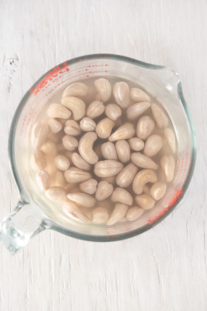 Cashews soaking in water in a large glass measuring cup, on a white wooden table top.