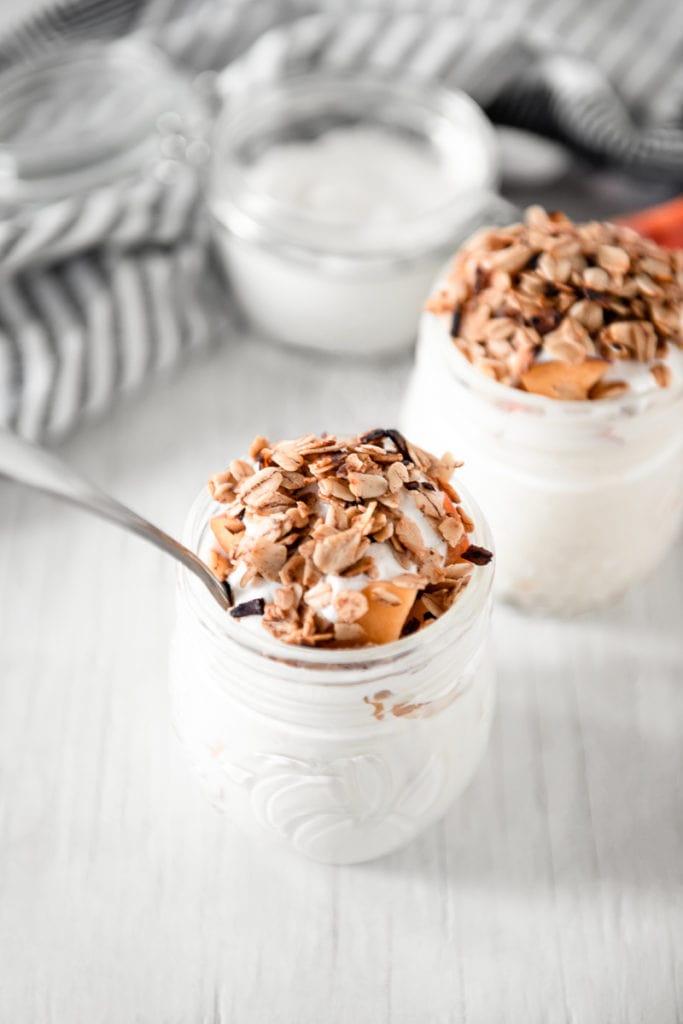 Peaches and cream topped with golden granola and cubed peaches in two round jam jars, the centre jar with a silver spoon. A sliced peach and a jar of cream on a linen cloth in the background
