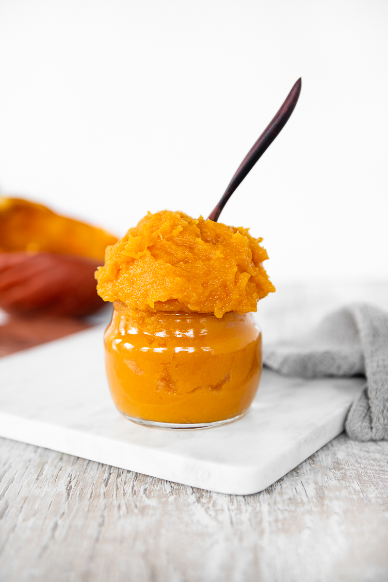 Homemade Pumpkin Purée heaping out of a glass jar with a wooden spoon sticking out the top.