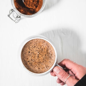 A hand grabbing a frothy french vanilla cappuccino and a jar of homemade french vanilla cappuccino mix with a spoon sticking out