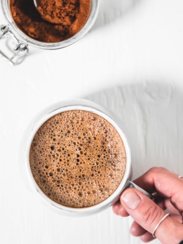 A hand grabbing a frothy french vanilla cappuccino and a jar of homemade french vanilla cappuccino mix with a spoon sticking out