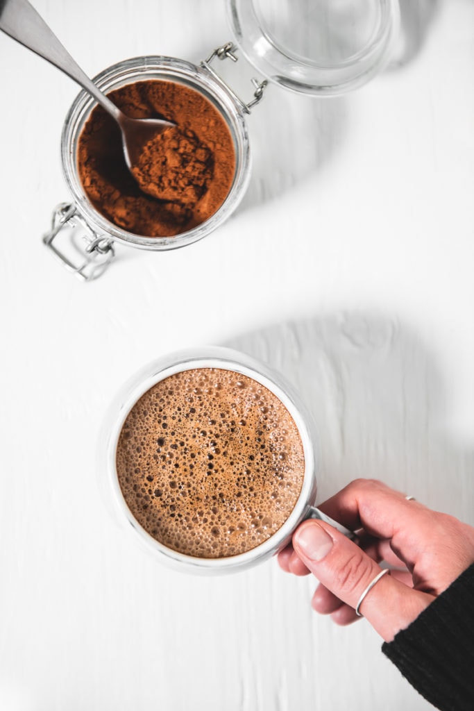 A hand lifting a frothy french vanilla cappuccino away from an open jar of instant mix.