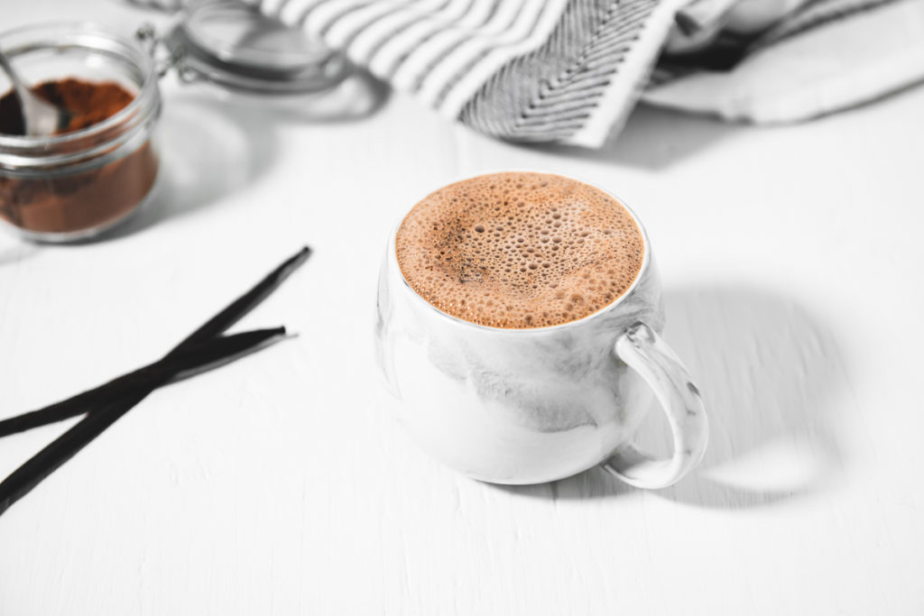 A grey and white marble mug filled with a frothy French Vanilla Cappuccino with two vanilla beans to the side a linen napkin and a spoon in a jar of Homemade French Vanilla Cappuccino Mix