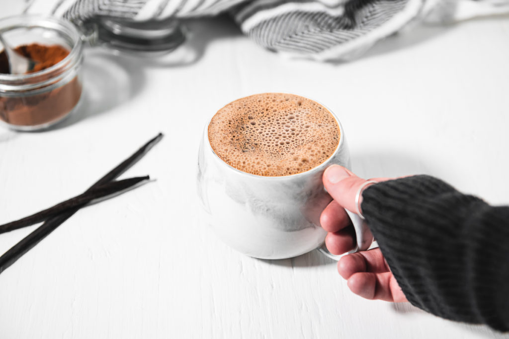 A hand grabbing a frothy french vanilla cappuccino, with two fresh vanilla beans and an open jar of Homemade Instant Cappuccino Powder.