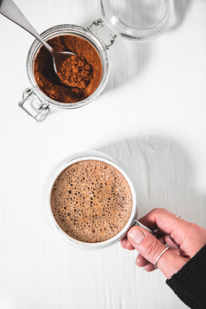 A hand grabbing a frothy french vanilla cappuccino and a jar of homemade french vanilla cappuccino mix with a spoon sticking out