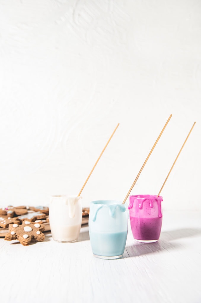 Three jars of naturally coloured icing with decorating sticks and icing dripping down the sides and a plate of gingerbread cookies beside.