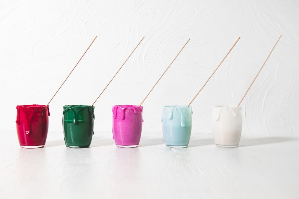 A row of homemade, naturally coloured icing (red, green, pink, blue and white) in jars with icing dripping down the sides and decorating sticks in each.