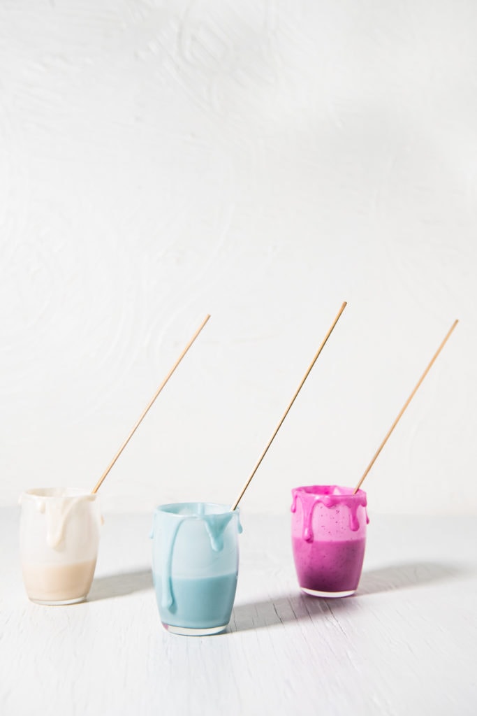 Three Jars of icing that's Naturally coloured (White, blue and pink) with icing dripping down the sides and a decorating stick in each jar.