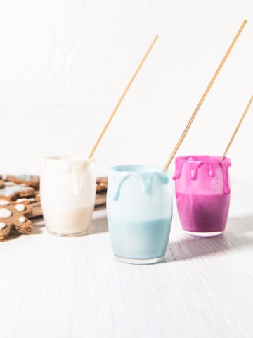 Three jars of naturally coloured icing with decorating sticks and icing dripping down the sides and a plate of gingerbread cookies beside.