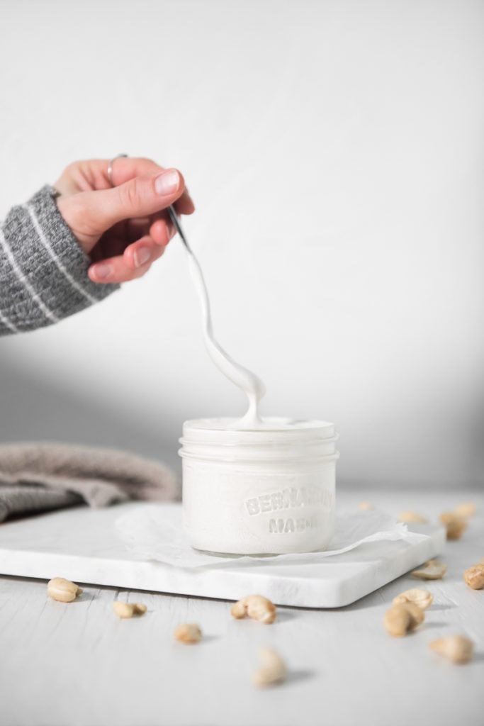A hand lifting a spoon out of a jar full of cashew cream. The cashew cream dripping off the spoon back into the jar.