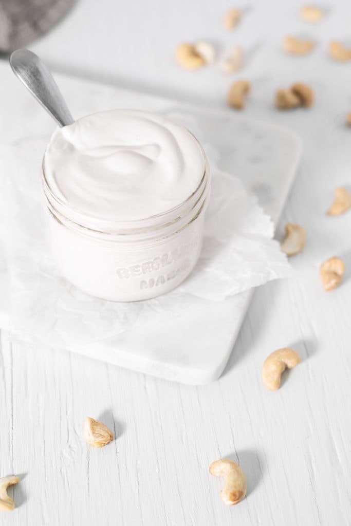 A hand lifting a dripping spoon out of a full jar of cashew cream beside a linen cloth and raw cashews sprinkled around.