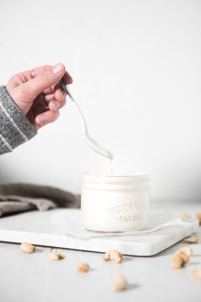 A hand lifting a dripping spoon out of a full jar of cashew cream beside a linen cloth and raw cashews sprinkled around.