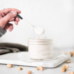 A hand dipping a spoon into a jar of cashew cream with raw cashews sprinkled around.