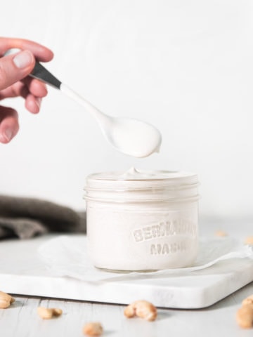 A hand dipping a spoon into a jar of cashew cream with raw cashews sprinkled around.