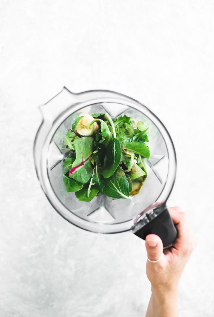 A hand grabbing the handle of a blender jug filled with fresh greens, banana, mooring powder, avocado and non-dairy milk