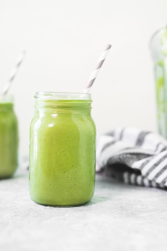 A green smoothie in a mason jar with a straw, another green smoothie behind with a linen cloth and an emptied blender jug on the other side.