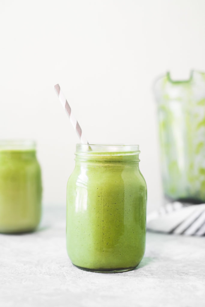 A mason jar filled with a vibrant green smoothie placed front and centre in front of another green smoothie, a white and grey striped linen cloth and an emptied blender jug.