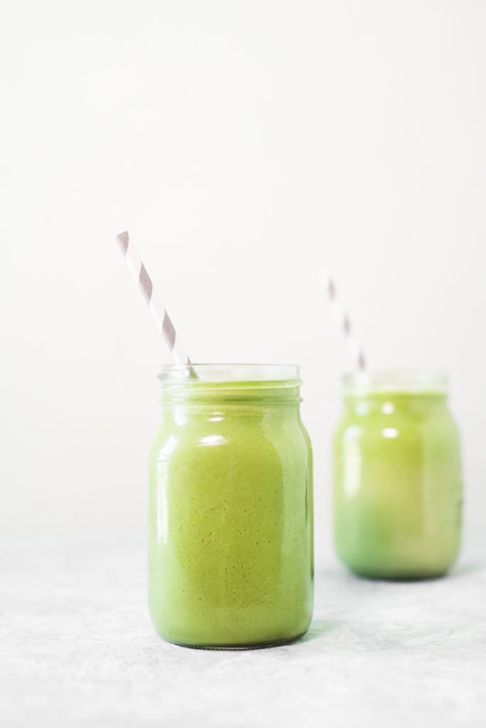 Two green smoothies in mason jars with white and grey striped straws sitting in front an empty blender Jug and a white and grey striped linen cloth.