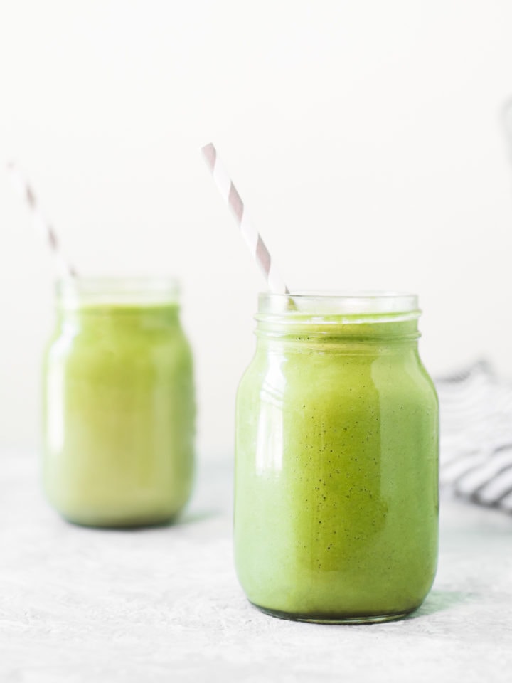 Two vibrant green avocado smoothies sitting beside a striped linen cloth and an empty blender jug. The smoothies also have white and grey striped straws perfectly placed, both pointing to the left.