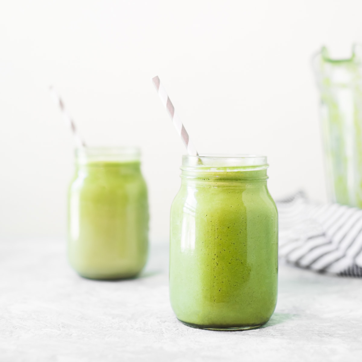 Two vibrant green avocado smoothies sitting beside a striped linen cloth and an empty blender jug. The smoothies also have white and grey striped straws perfectly placed, both pointing to the left.