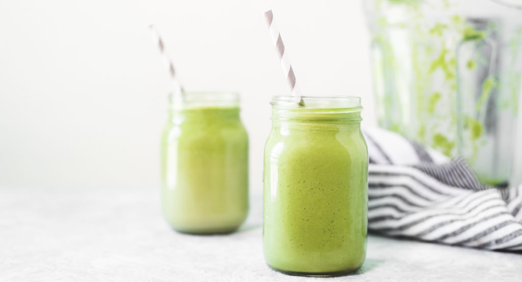 Two vibrant green avocado smoothies sitting beside a striped linen cloth and an empty blender jug. The smoothies also have white and grey striped straws perfectly placed, both pointing to the left.