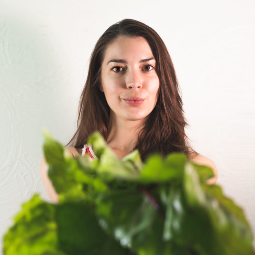 A smiling woman with long dark hair holding a bunch of beet greens toward the camera.