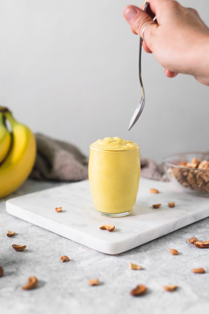 A little glass pudding cup full of banana turmeric pudding with a hand holding a spoon over top about to take a scoop.