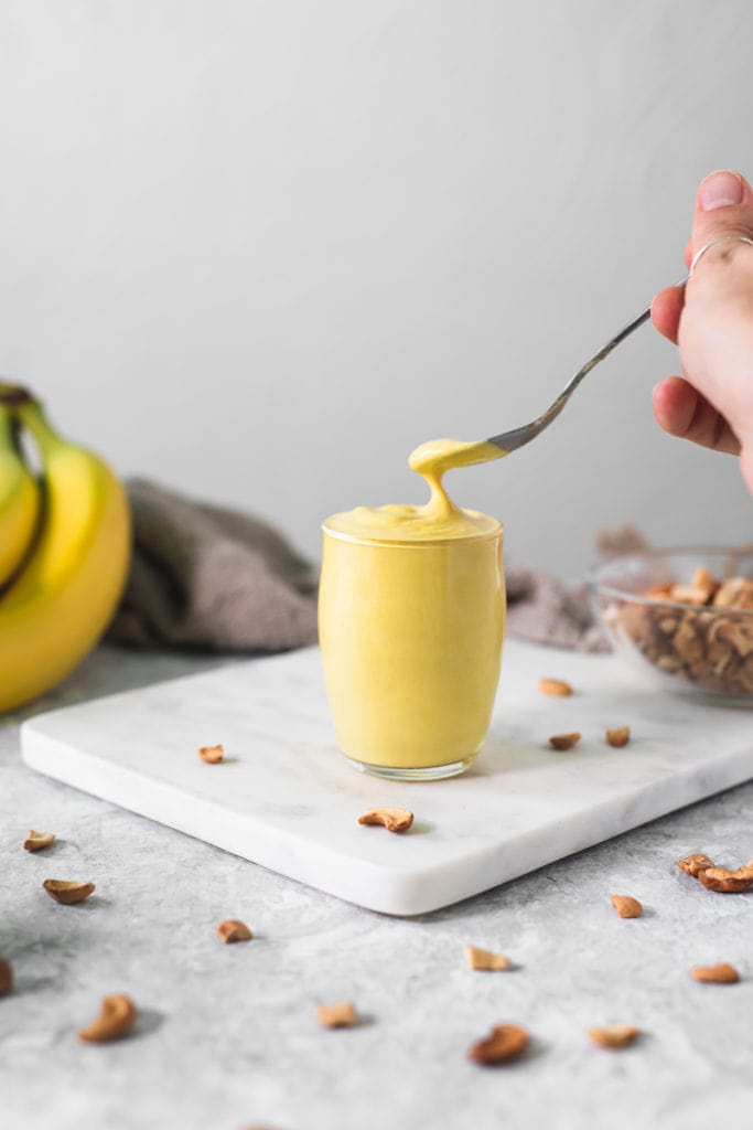 Creamy Banana Pudding being scooped out of a little glass cup, surrounded by toasted cashews, a bunch of bananas and a linen cloth.