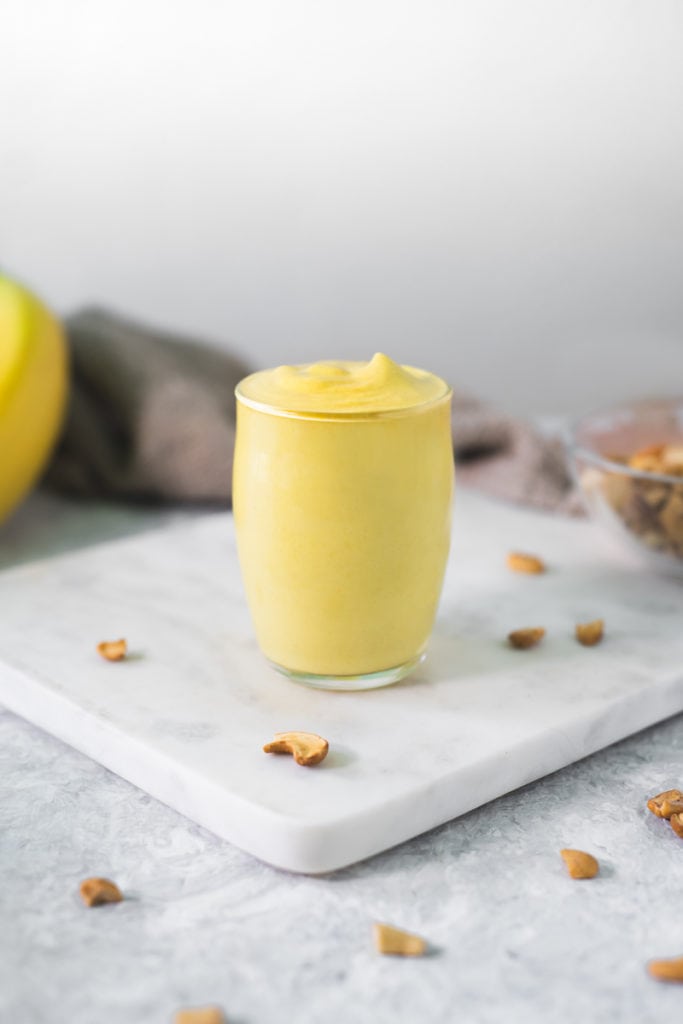 A full cup of healthy vegan banana cream pudding surrounded by toasted cashews, a bunch of bananas and grey linen cloth.