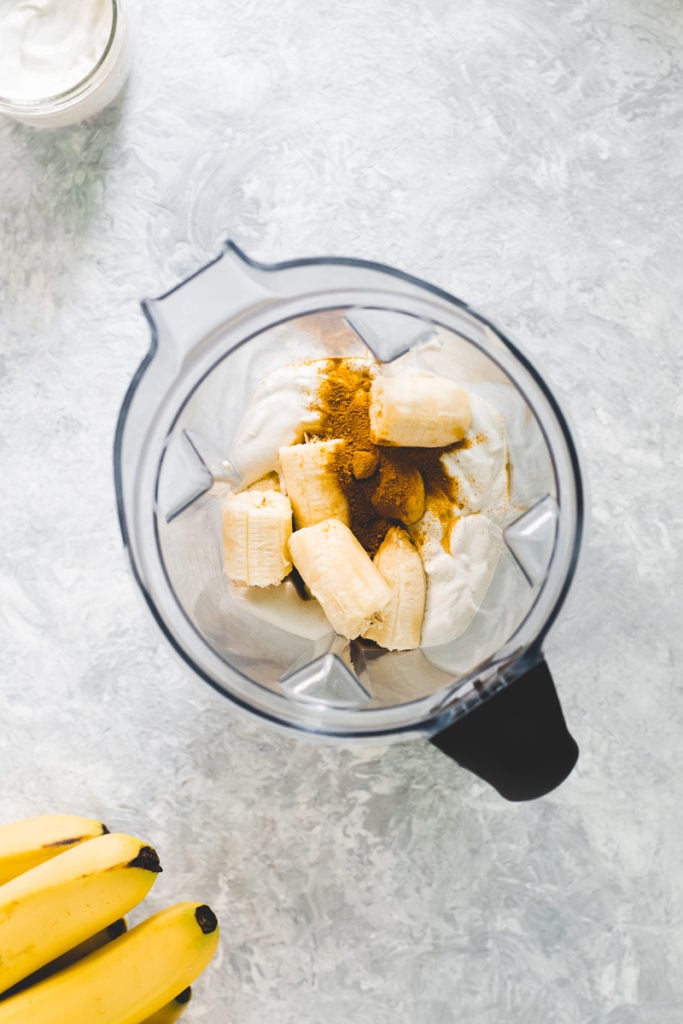 A blender jug filled with banana, turmeric powder, vanilla extract and cashew cream.