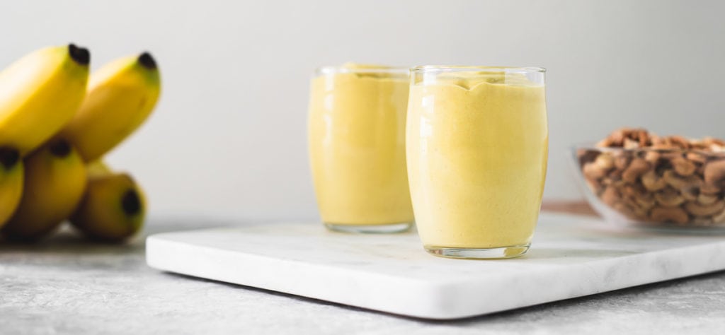 Two cups of yellow banana cream pudding sitting on a marble cutting board in between a bowl of toasted cashews and a bunch of bananas.