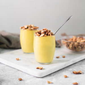 Two cups of banana yellow pudding topped with toasted cashews. The pudding cups are surrounded by dropped cashews pieces, a bowl of cashews and a linen cloth.