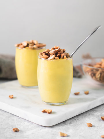 Two cups of banana yellow pudding topped with toasted cashews. The pudding cups are surrounded by dropped cashews pieces, a bowl of cashews and a linen cloth.