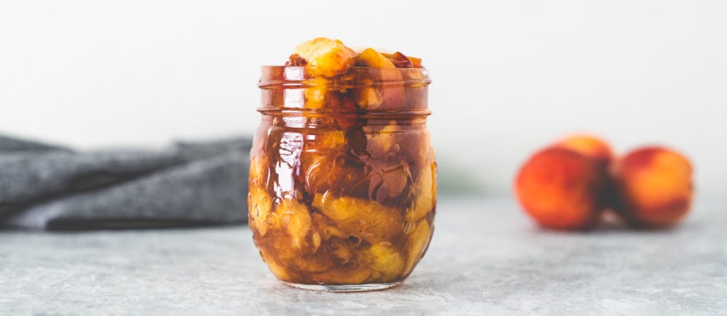 A heaping jar of fresh peach pie filling sitting centred in front of a bunch of peaches and a grey linen cloth.