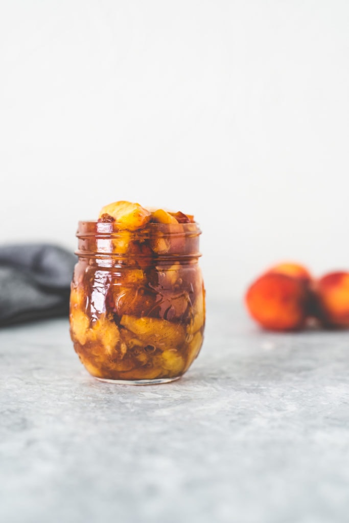 A jar full of fresh peach pie filling sitting in front of a bunch of peaches and a grey linen cloth.